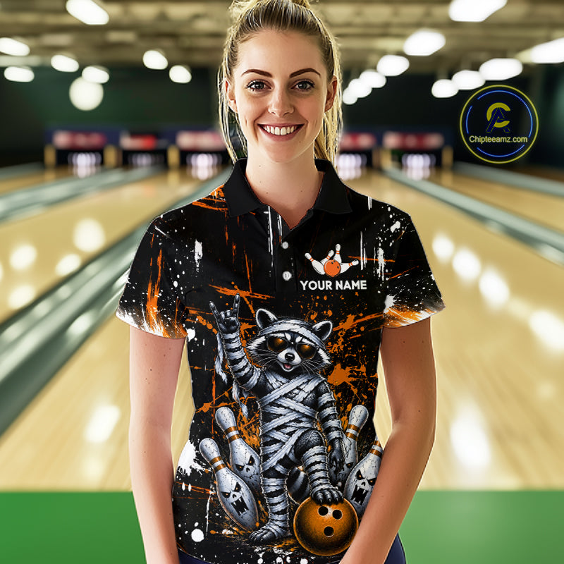 A woman stands in front of a bowling alley wearing a black short-sleeve polo shirt with a raccoon and bowling ball graphic, orange paint splatters, white bowling pins, and the text 'YOUR NAME' on the chest. 