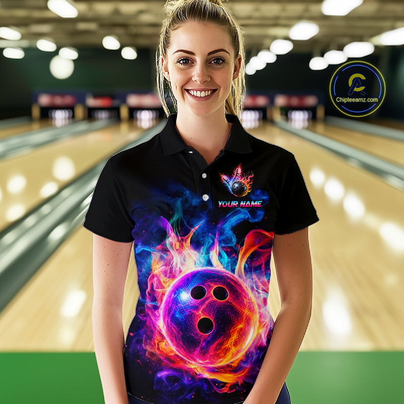 A woman stands in a bowling alley wearing a black short-sleeve polo shirt with a vibrant, colorful graphic of a glowing bowling ball surrounded by flames. The shirt features a custom name placeholder and a circular logo on the left chest.