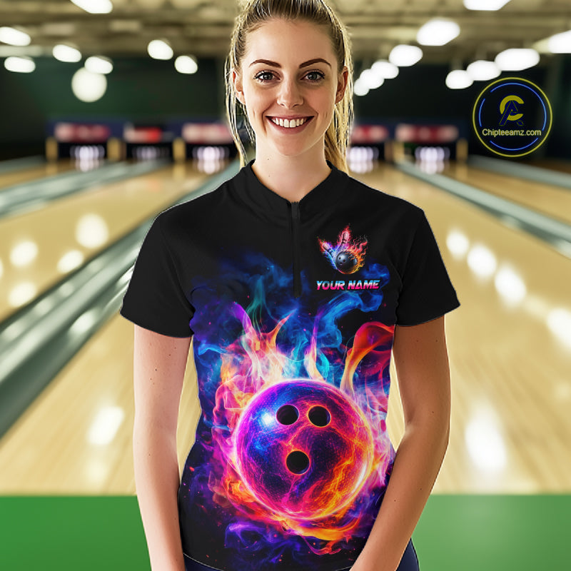 A woman stands in a bowling alley wearing a black short-sleeve polo shirt with a vibrant, colorful graphic of a flaming bowling ball and the text 'YOUR NAME'. 