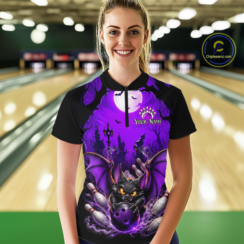 A woman wearing a black and purple Halloween-themed bowling bowling shirt stands in a bowling alley. The shirt features a graphic of a black bat with glowing yellow eyes, bowling pins, and a full moon under a purple sky, with 'Your Name' printed on the chest.