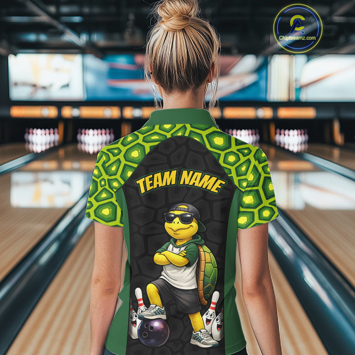 A woman viewed from behind wearing a green and black short-sleeve bowling-themed quarter-zip shirt with a cartoon turtle graphic and 'TEAM NAME' text on the back, standing in a bowling alley.