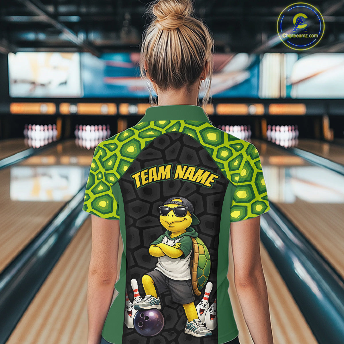 A person viewed from behind wearing a short-sleeve bowling shirt in a bowling alley. The shirt has a green and black turtle-scale pattern, yellow 'TEAM NAME' text, and a cartoon turtle graphic on the back.