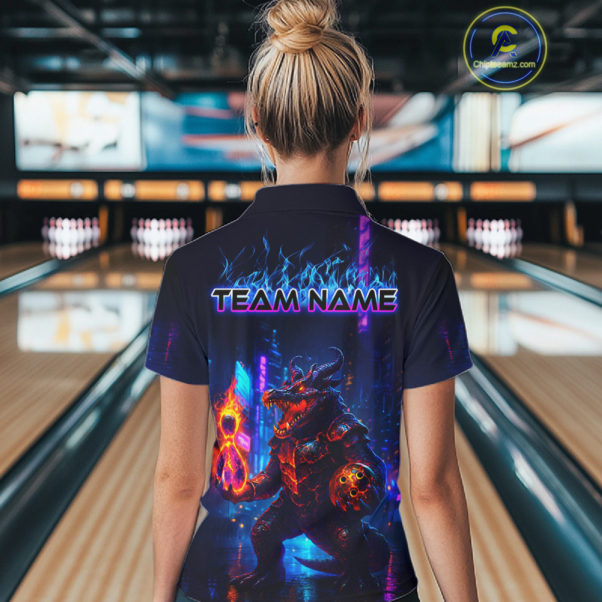 A woman viewed from behind wearing a dark navy short-sleeve shirt with a large colorful bowling-themed graphic on the back and the text 'TEAM NAME' across the shoulders. The background shows a blurred bowling alley with lanes and pins.