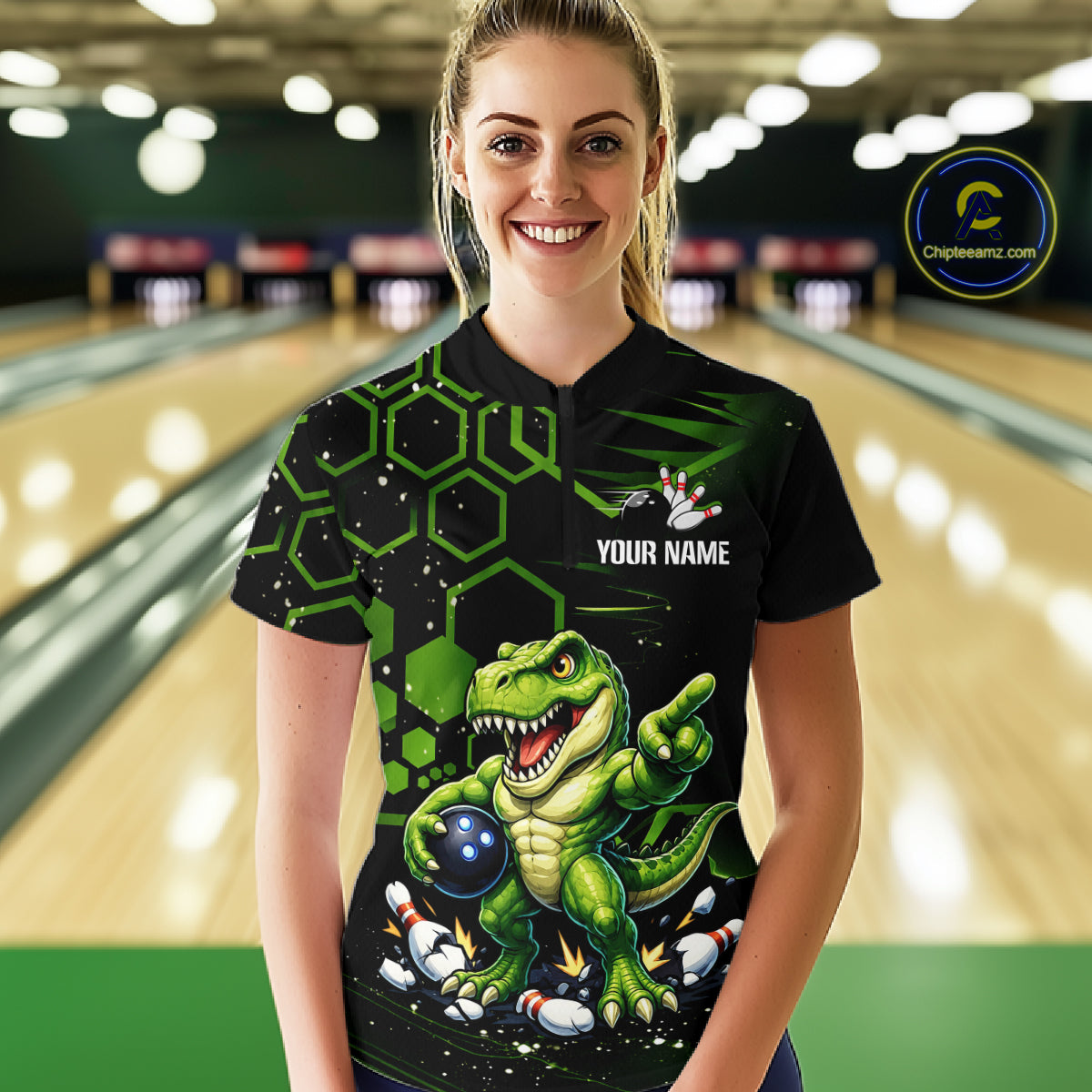 A woman stands in a bowling alley wearing a black short-sleeve bowling shirt with a green cartoon dinosaur graphic and the text 'YOUR NAME'. The background shows blurred bowling lanes, and a circular 'Championship.