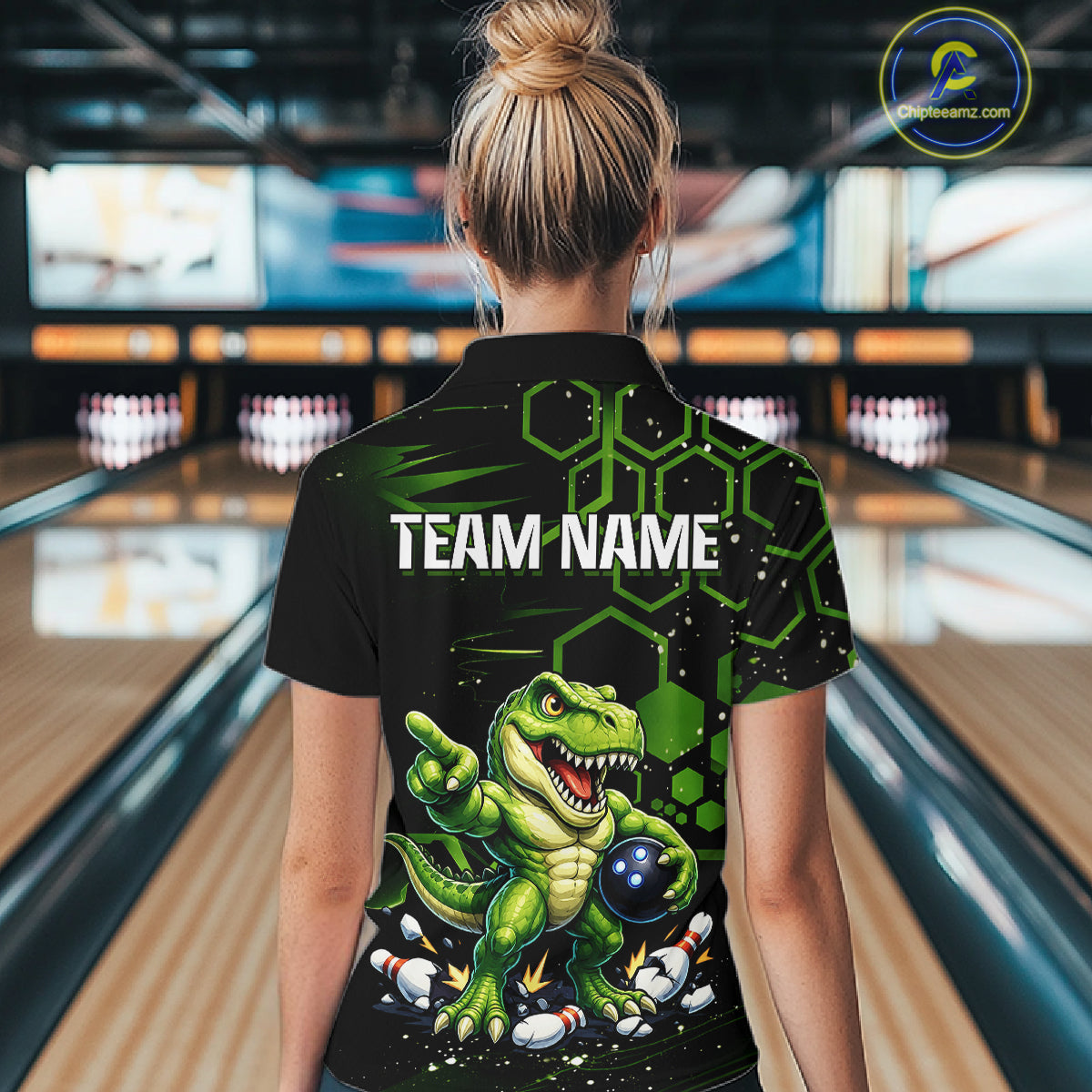 A woman viewed from behind wearing a black short-sleeve bowling shirt with a green cartoon dinosaur graphic and the text 'TEAM NAME' on the back, standing in a bowling alley.