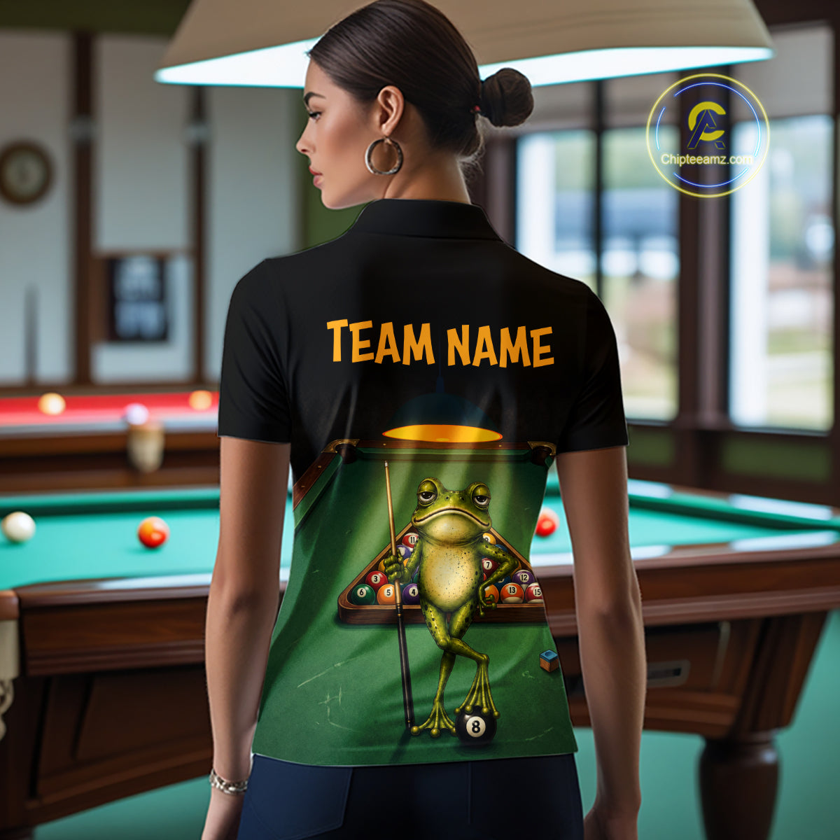 A woman stands in a billiards room wearing a black short-sleeve polo shirt from the back. The shirt features a green graphic of a frog playing pool with the text 'TEAM NAME' in yellow.