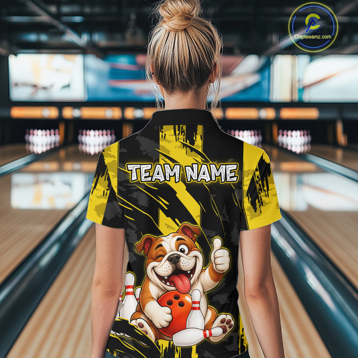 Female model wearing a custom black yellow bulldog bowling polo shirt back view displaying the personalized team name above a playful cartoon bulldog graphic at the bowling alley