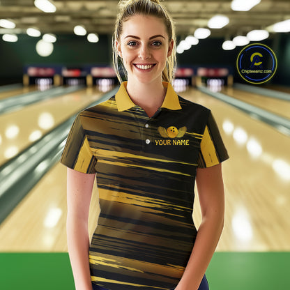 Female model wearing a custom black gold bowling polo shirt front view featuring a gold bowling ball emblem, dynamic gold brush stroke pattern and striped design at the bowling alley