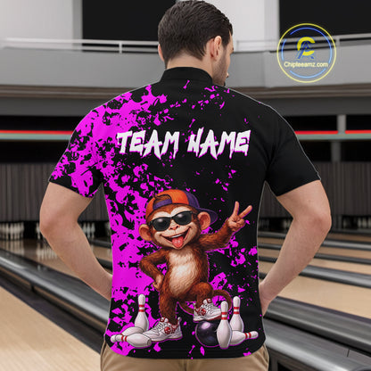 A man viewed from behind wearing a black short-sleeve bowling shirt with a bright purple splatter design, white 'TEAM NAME' text, and a cartoon monkey graphic on the back. Bowling lanes are visible in the background.
