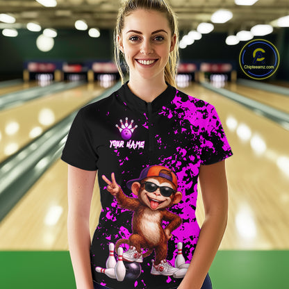 A woman standing in a bowling alley wearing a black short-sleeve bowling shirt with a bright purple paint splatter design, a cartoon monkey graphic, and a purple bowling pin crown logo. The background shows blurred bowling lanes.