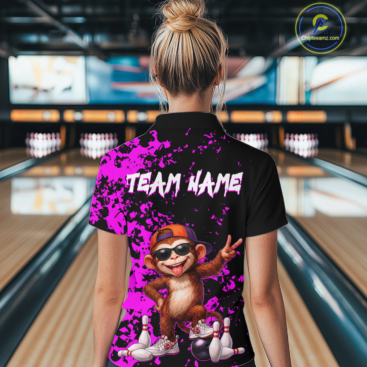 A woman viewed from behind wearing a black short-sleeve polo shirt with a bright purple splatter design, white 'TEAM NAME' text, and a cartoon monkey graphic in bowling lanes. The image is set in a bowling alley.