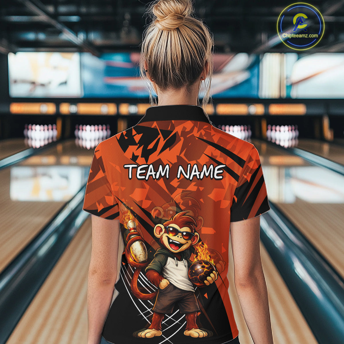 A woman viewed from behind wearing a short-sleeve bowling-themed polo shirt in a bowling alley. The shirt features a bold orange and black abstract pattern, the text 'TEAM NAME' across the upper back, and a cartoon monkey holding bowling balls near the lower back.