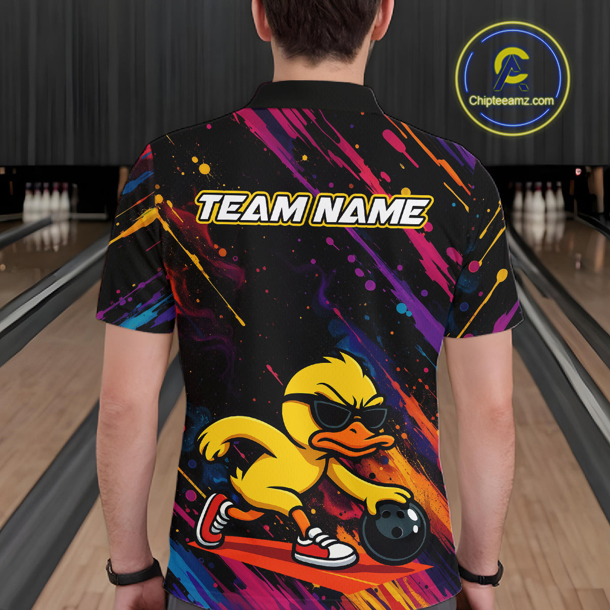 A person viewed from behind wearing a black short-sleeve custom bowling shirt with a colorful paint-splatter design, a yellow cartoon duck wearing sunglasses, and the text 'TEAM NAME' across the back. The background shows bowling lanes in a dimly lit bowling alley.