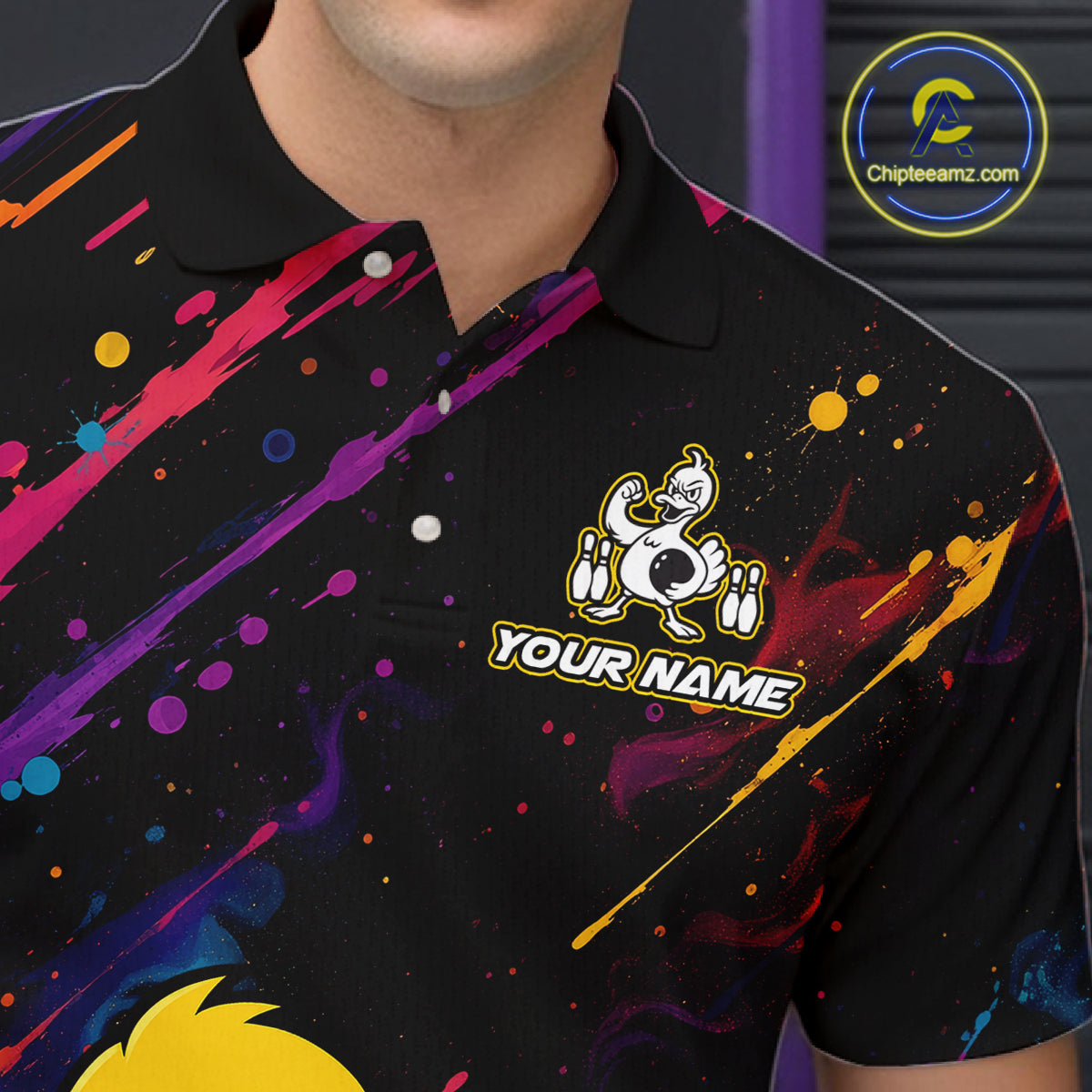 A close-up of a person wearing a black short-sleeve polo shirt with colorful paint-splatter graphics in purple, pink, blue, yellow, and orange. The shirt features a cartoon duck logo with the text 'YOUR NAME' on the chest.