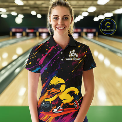 A woman stands in a bowling alley wearing a black short-sleeve polo shirt with a colorful graphic of a yellow duck bowling. The shirt features a galaxy print on the upper chest and a large yellow duck illustration with sunglasses, a bowling ball, and the text 'YOUR NAME' on the front.