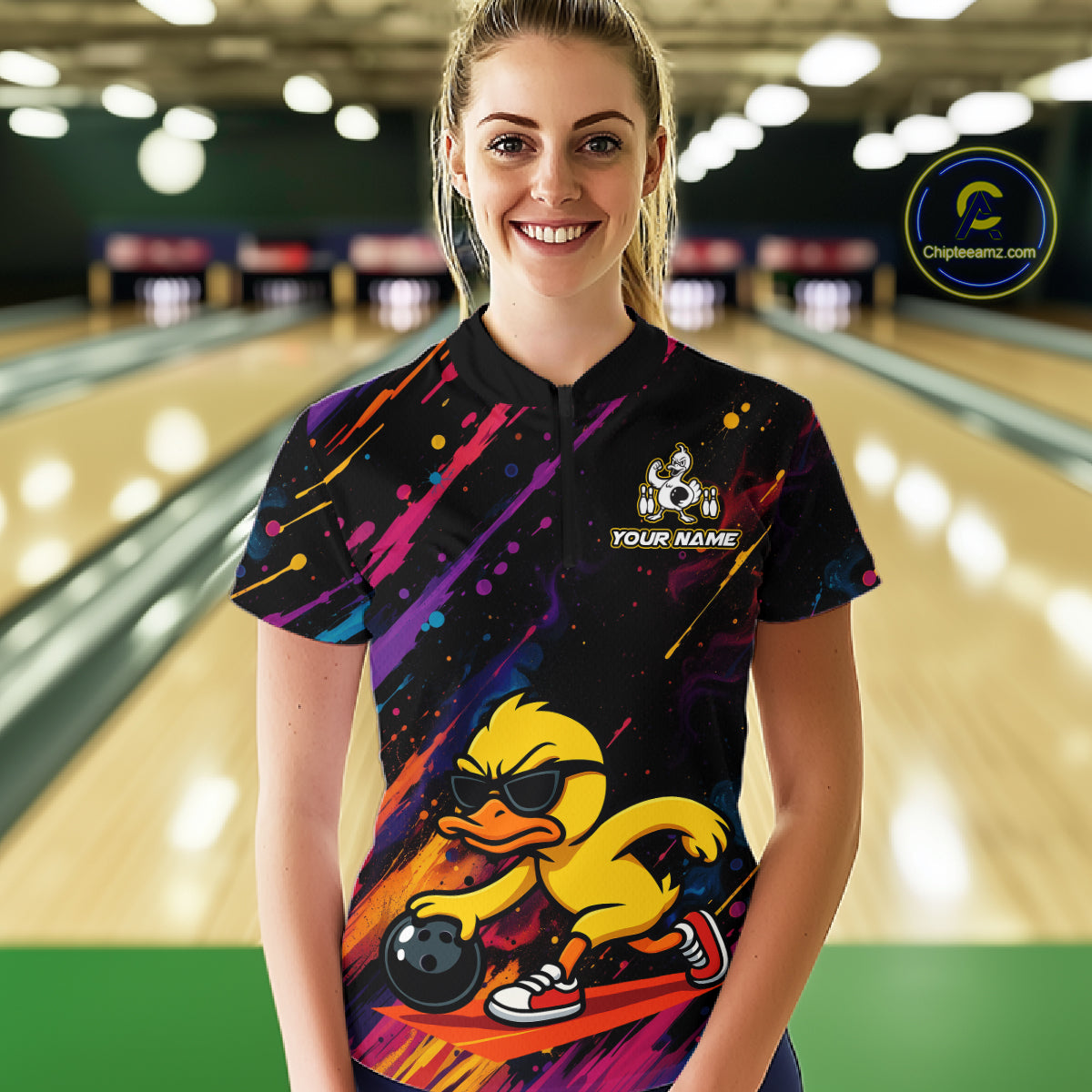 A woman stands in a bowling alley wearing a black short-sleeve bowling shirt with a colorful paint-splatter design and a large yellow duck graphic on the front. The shirt has a white cartoon duck logo and the text 'YOUR NAME' on the chest.