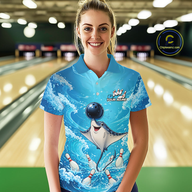 A woman wearing a bright blue short-sleeve bowling-themed polo shirt with a cartoon stingray and bowling pins graphic, standing in a bowling alley with blurred lanes in the background.