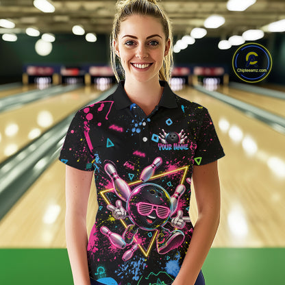 A woman stands in a bowling alley wearing a black short-sleeve polo shirt with a colorful retro bowling-themed graphic print featuring bowling pins, balls, and neon splatter details. The background shows blurred bowling lanes and lights.