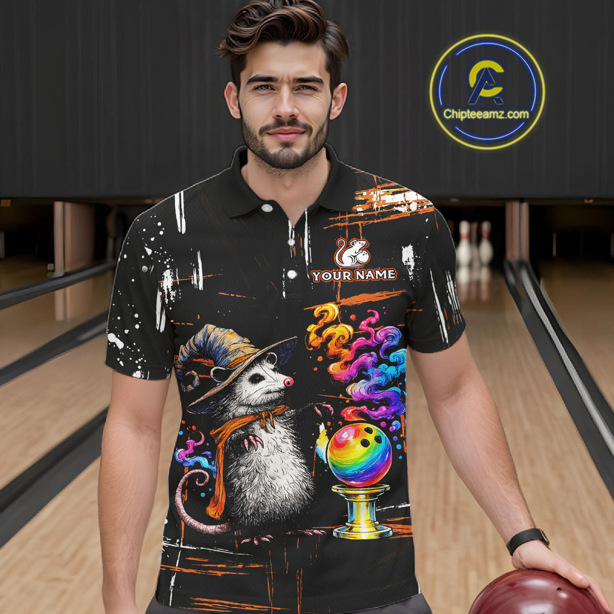 A man wearing a black short-sleeve bowling-themed graphic polo shirt with a colorful cartoon opossum, bowling ball, and spray-paint splatter design, standing in a bowling alley. 
