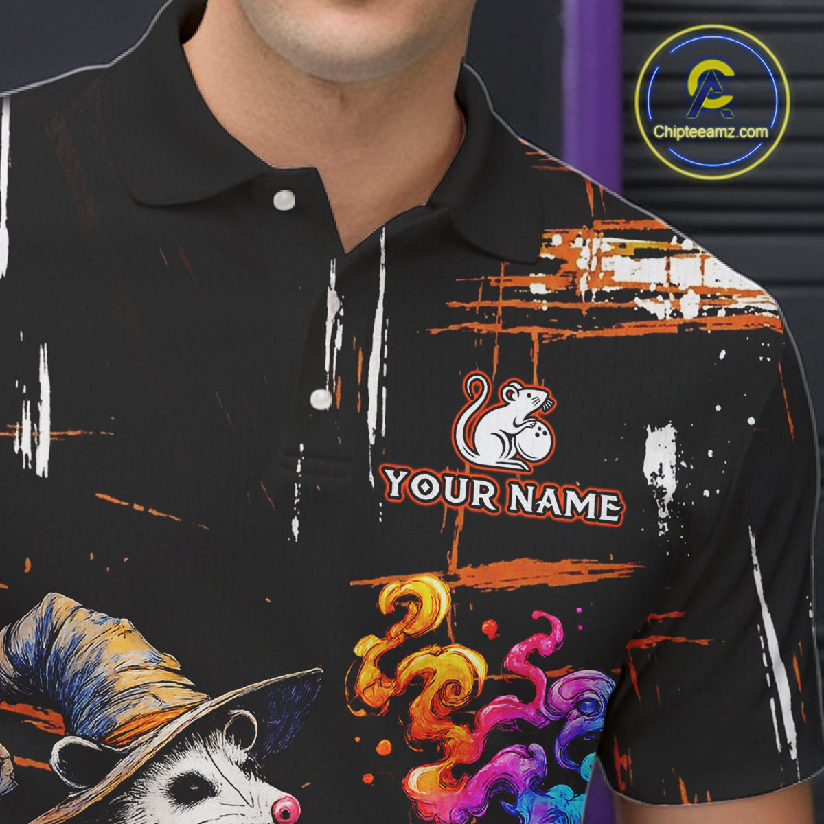 A close-up of a black short-sleeve polo shirt with a colorful custom graphic print featuring a mouse illustration, paint splatters, and a placeholder logo reading 'YOUR NAME'.