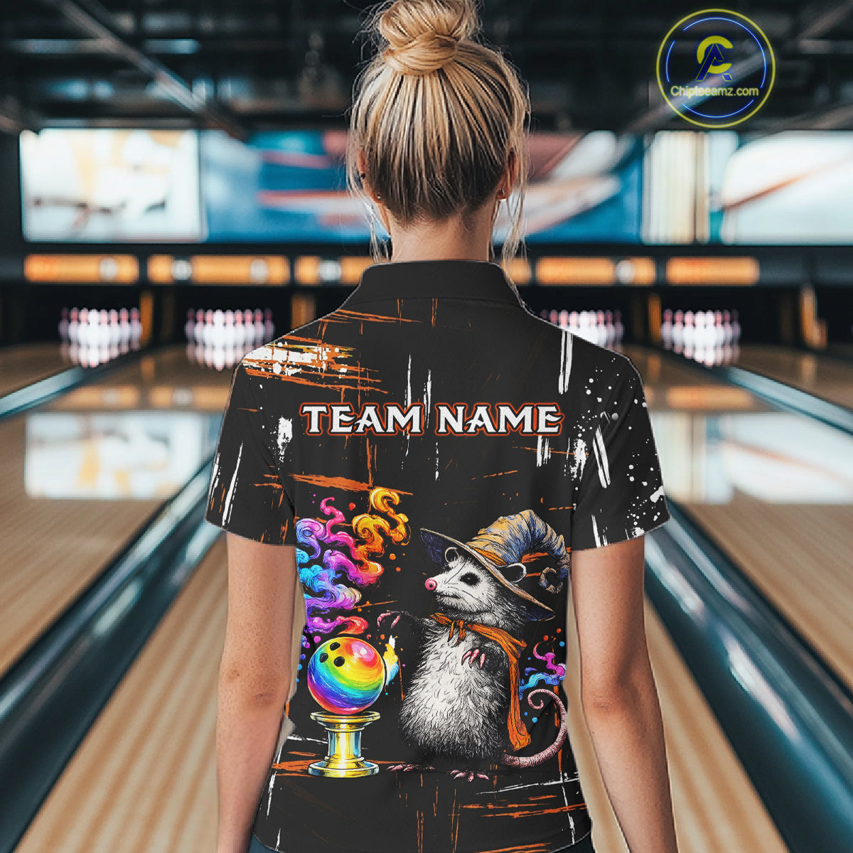 A woman viewed from behind wearing a black short-sleeve bowling-themed graphic shirt in a bowling alley. The shirt features a large printed design with the text 'TEAM NAME', a raccoon in a hat, a bowling ball, and colorful splatter details.