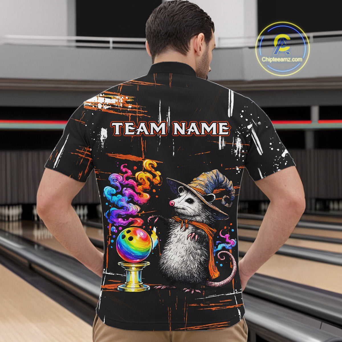 A man viewed from behind wearing a black bowling-themed short-sleeve shirt with a colorful graphic of a possum in a witch hat next to a rainbow bowling ball, with 'TEAM NAME' printed across the upper back. The background shows bowling lanes.