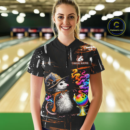 A woman wearing a black short-sleeve bowling-themed t-shirt with a colorful cartoon opossum, bowling ball, and paint splatter design, standing in front of a blurred bowling alley background. 