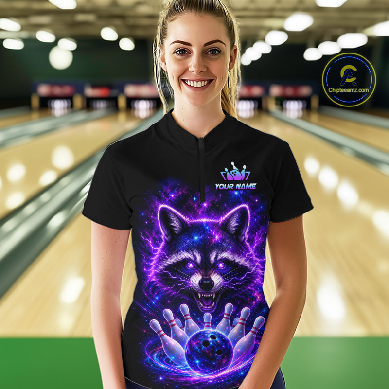 A woman stands in a bowling alley wearing a black short-sleeve bowling shirt with a large purple and blue cosmic raccoon graphic on the front. The shirt has a mock logo and the text 'YOUR NAME'.