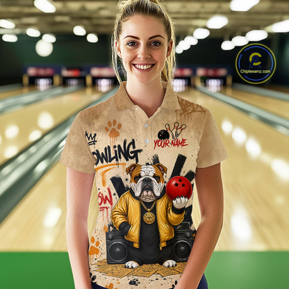A woman standing in a bowling alley wearing a beige short-sleeve button-up shirt with a custom bulldog bowling graphic. The shirt features a bulldog in a yellow jacket holding a red bowling ball, with 'BOWLING' and 'YOUR NAME' text.
