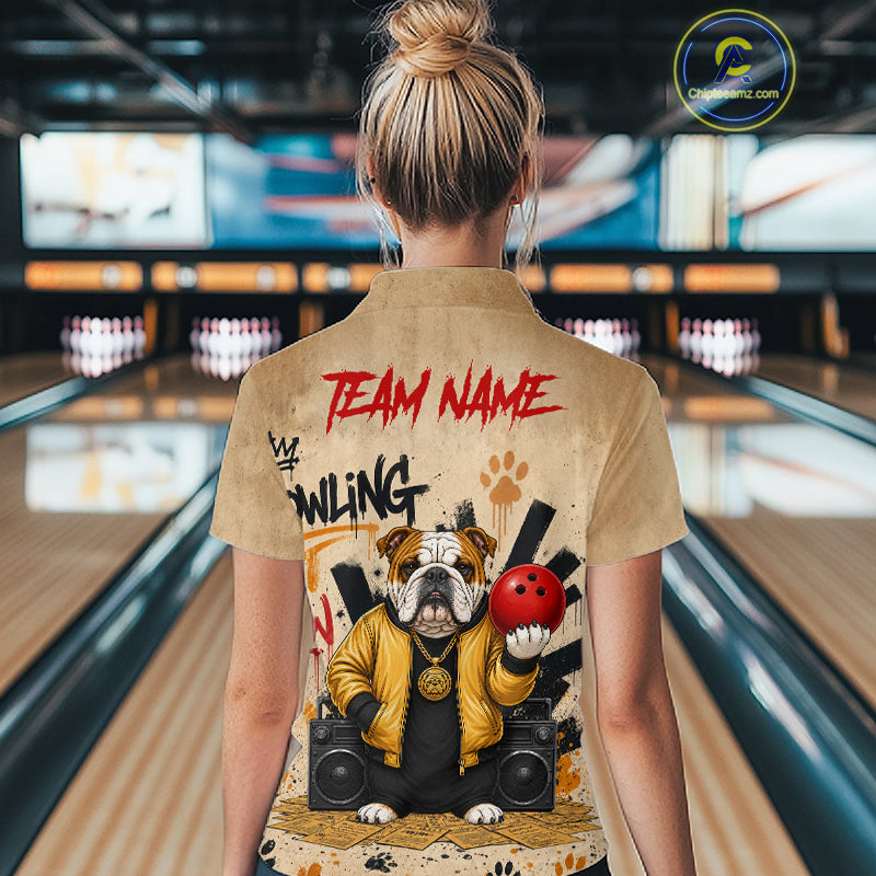 A woman viewed from behind wearing a tan short-sleeve bowling shirt with a large graphic print of a bulldog holding a red bowling ball and the words 'TEAM NAME' and 'BOWLING'. The background shows a blurred bowling alley with lanes and pins.