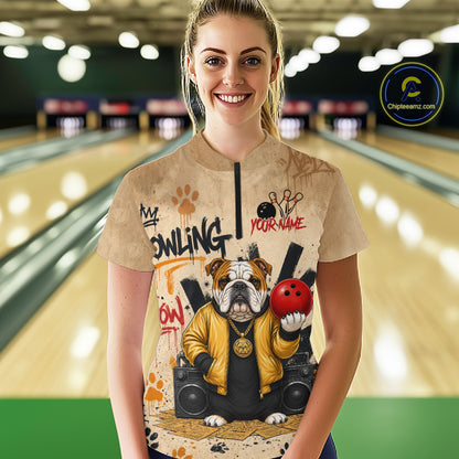 A woman stands in a bowling alley wearing a beige graphic t-shirt featuring a bulldog holding a red bowling ball with the words 'MY BOWLING YOUR NAME'. The background shows blurred bowling lanes, and a 'Chilewich.