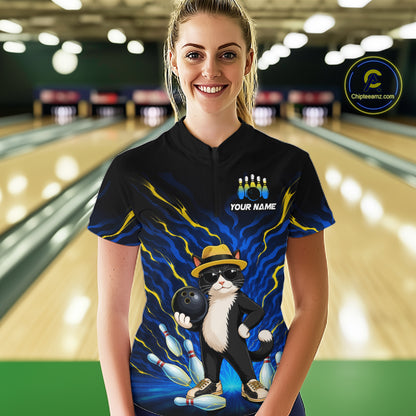 A woman stands in a bowling alley wearing a black short-sleeve bowling t-shirt with a colorful graphic of a cat bowling and the text 'YOUR NAME'. The background shows blurred bowling lanes.