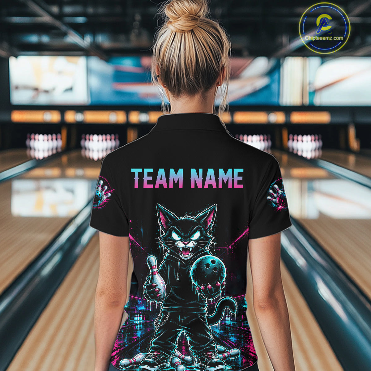 A woman viewed from behind wearing a black short-sleeve polo shirt with a large colorful graphic of a cartoon cat holding a bowling ball on the back, with 'TEAM NAME' printed above it. The background shows blurred bowling alley lanes.