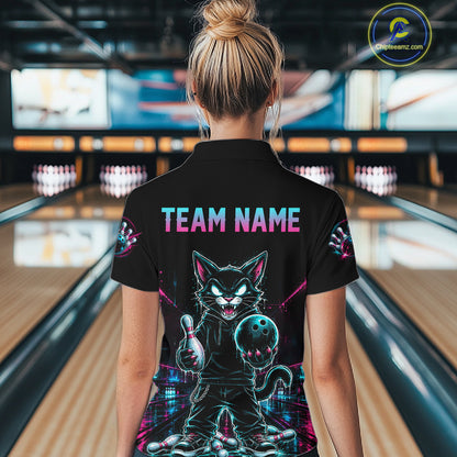 A woman viewed from behind wearing a black short-sleeve polo shirt with a large colorful graphic of a cartoon cat holding a bowling ball on the back, with 'TEAM NAME' printed above it. The background shows blurred bowling alley lanes.