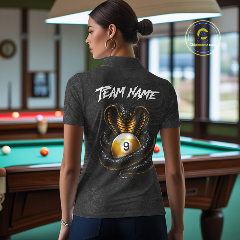 A woman stands in a billiards room wearing a dark gray short-sleeve polo shirt from the back, featuring a graphic of a gold 9-ball with a coiled black and gold snake and the text 'TEAM NAME' in white. A circular 'CheekyWear.
