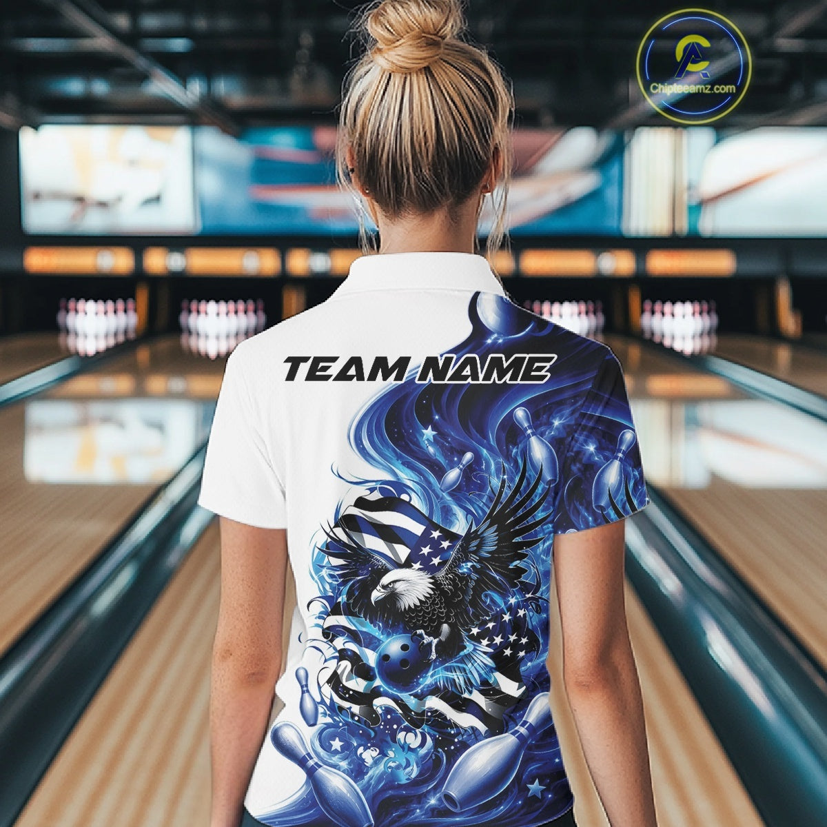 Back view female model at a bowling lane wearing a white and blue polo shirt with eagle graphics, bowling elements, and flowing flag inspired artwork.