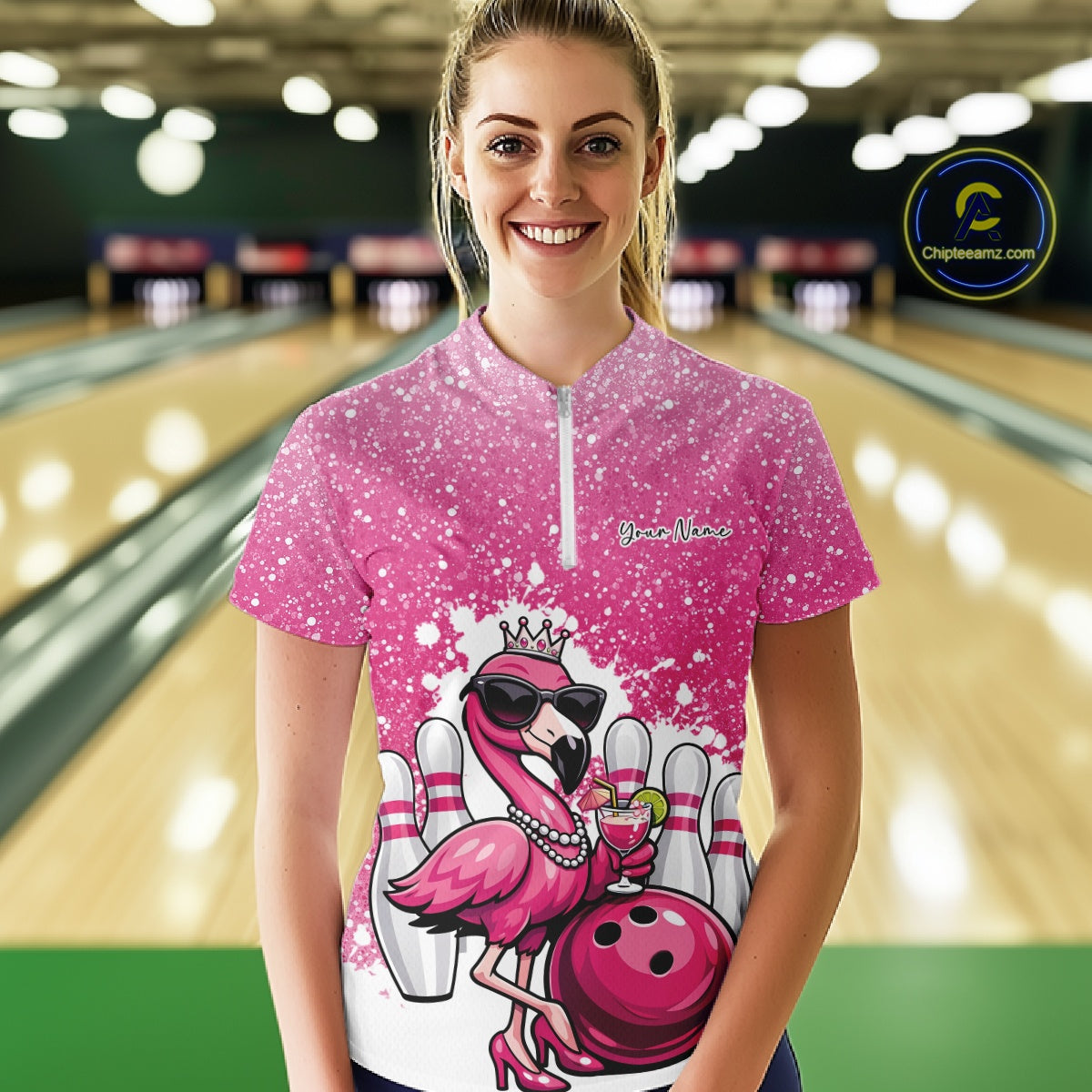 Female model at a bowling lane wearing a pink bowling quarter-zip shirt with a crowned flamingo, cocktail, bowling ball, and paint splatter graphics.