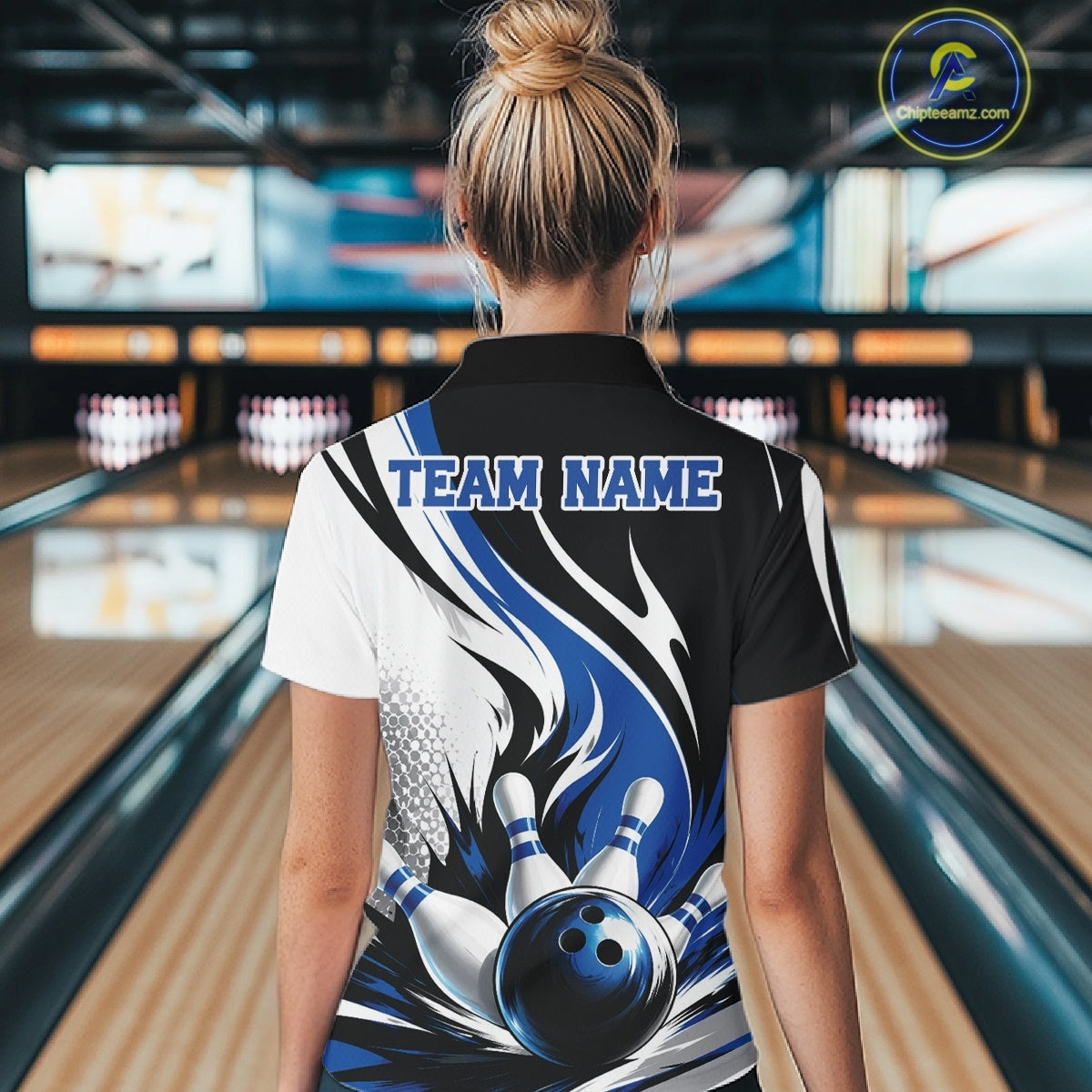 Back view of a female model wearing a white, black, and blue bowling polo shirt with a flaming bowling ball knocking down pins graphic at a bowling lane.