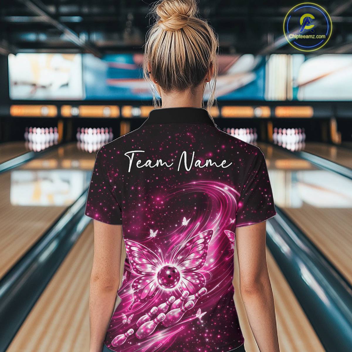 Back view of a female model at a bowling lane wearing a pink bowling quarter-zip shirt featuring glowing butterfly wings and galaxy-themed artwork.