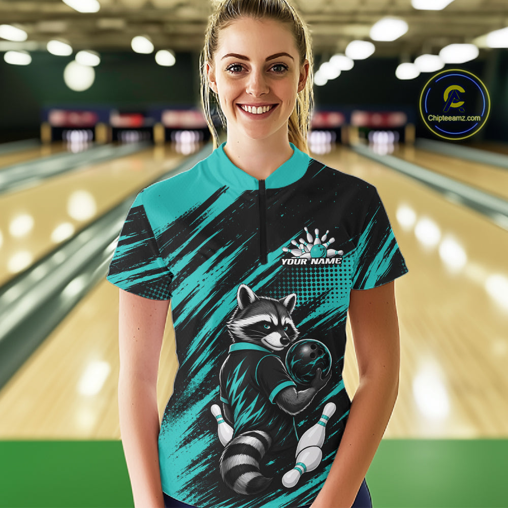 A woman stands in a bowling alley wearing a short sleeve bowling quarter zip shirt with a teal and black brushstroke design, a raccoon bowling graphic, and a mock logo on the chest. 