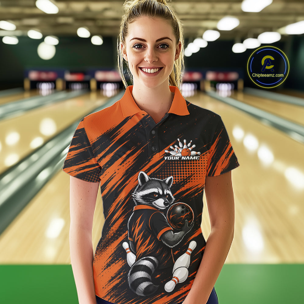 A woman stands in a bowling alley wearing a short-sleeve polo shirt with a bold orange and black brushstroke design, a raccoon holding a bowling ball graphic, and a white 'YOUR NAME' placeholder print on the chest. 
