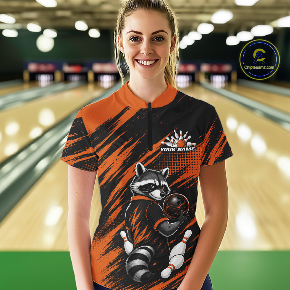 A woman stands in a bowling alley wearing a short-sleeve bowling shirt with a raccoon graphic. The shirt features a black and orange brushstroke pattern with a raccoon holding a bowling ball
