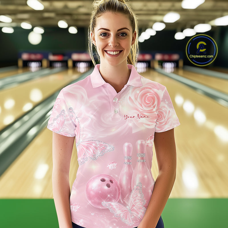 Female model at a bowling lane wearing a bowling polo shirt with pink glitter butterfly artwork, roses, bowling pins, and a bowling ball on the front.