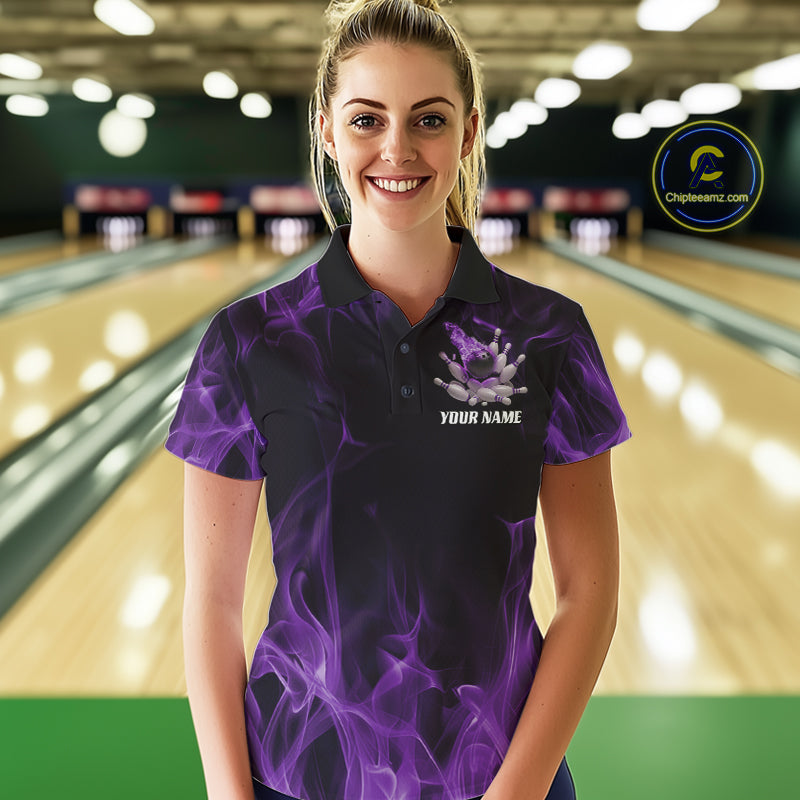 Female model at a bowling lane wearing a black and purple polo shirt featuring a dynamic bowling ball crash graphic with flames and scattered pins.