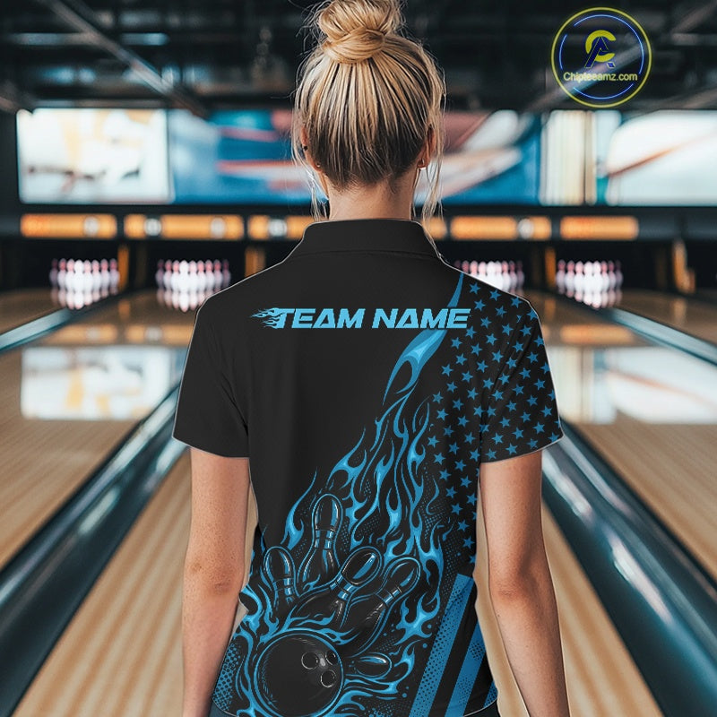 Back view of a female model in a bowling center wearing a black polo shirt featuring a blue flaming bowling ball knocking down pins.