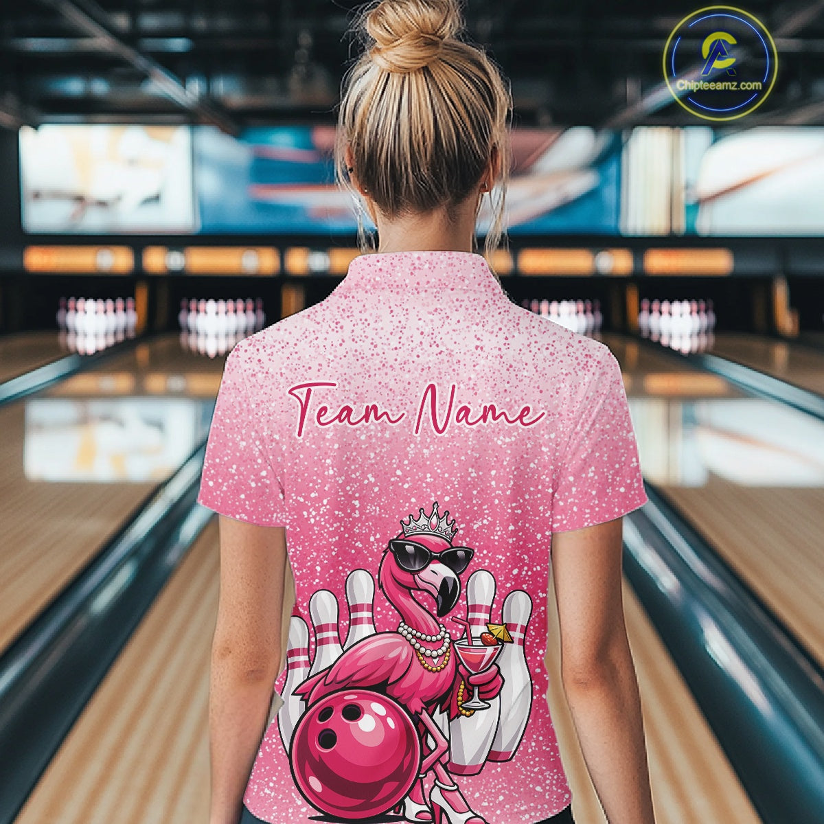 Back view of a female model in a bowling alley wearing a pink quarter-zip bowling shirt showcasing crowned flamingo and bowling-themed artwork.