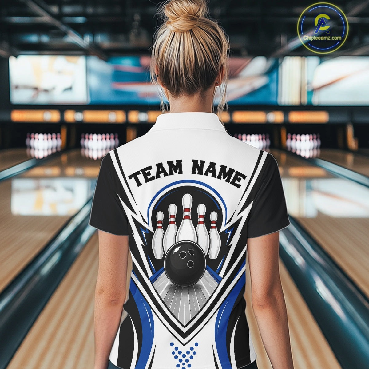 Female model shown from the back wearing a white, black, and blue bowling polo shirt with bowling ball striking pins artwork at a bowling lane.