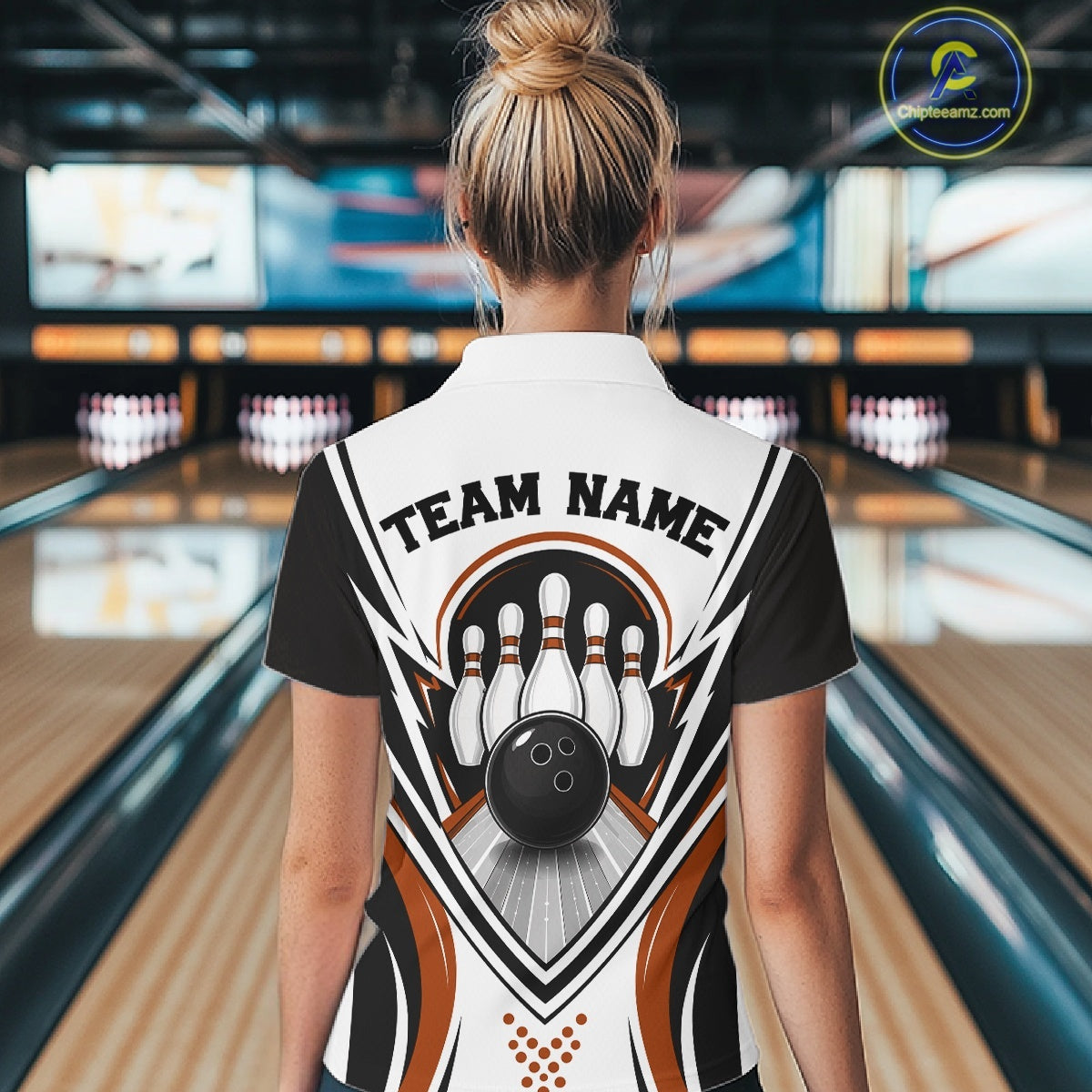 Back view of a female model inside a bowling center wearing a white, black, and orange bowling polo shirt featuring bowling ball striking pins graphics.