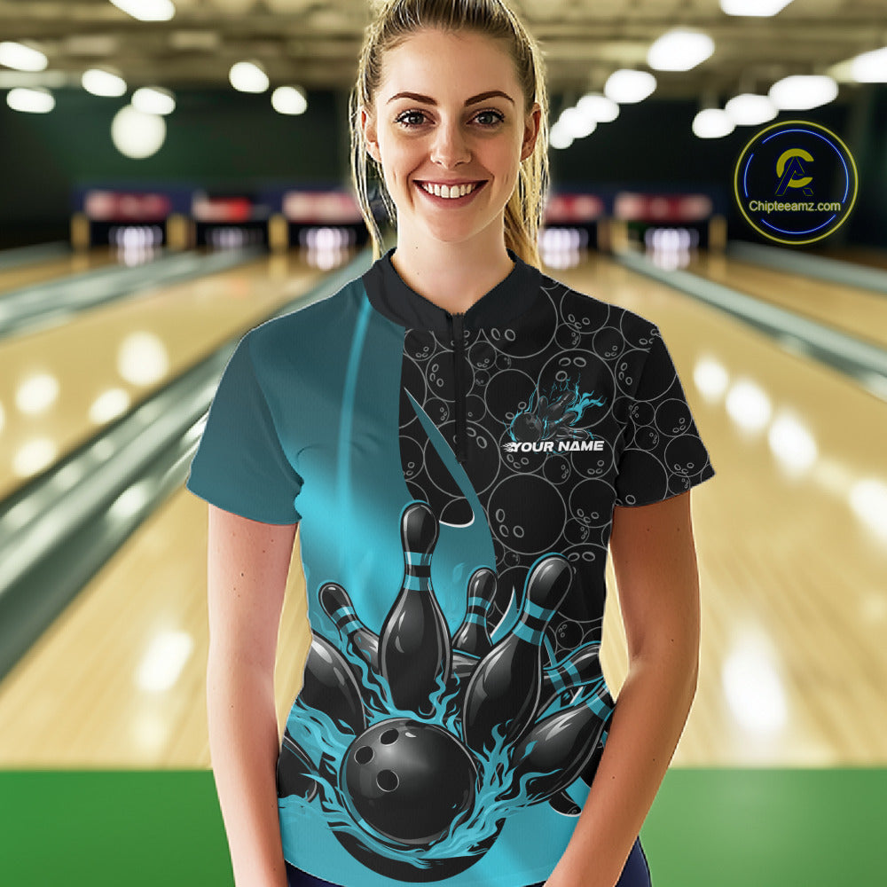 Front view of a female model wearing a black and blue bowling quarter-zip shirt at a bowling lane, featuring flaming bowling ball striking pins artwork with gradient ball pattern graphics.
