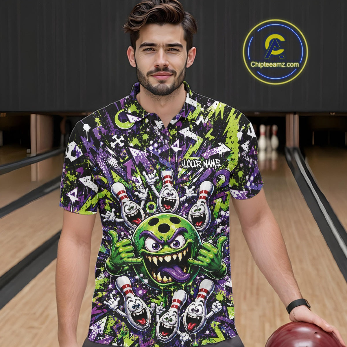 Front view of a male model at a bowling lane wearing a green and purple bowling polo shirt featuring a mischievous green monster bowling ball with sharp teeth, a purple tongue, playful hand gesture, and cheerful cartoon bowling pins.